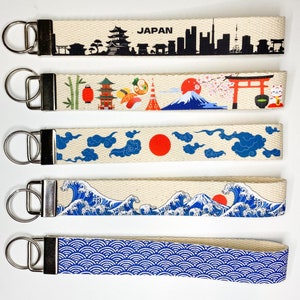 May include: Five different keychains with a white background and various Japanese-themed designs. The designs include a cityscape, a mountain with cherry blossoms, clouds with a red sun, waves, and a blue and white pattern.