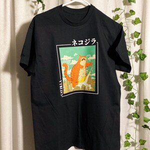 May include: Black t-shirt with a graphic print of a cartoon orange catzilla monster with the text "CATZILLA" and "ネコジラ" in Japanese.