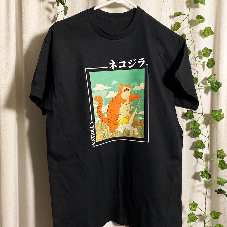 May include: Black t-shirt with a graphic print of a cartoon orange catzilla monster with the text "CATZILLA" and "ネコジラ" in Japanese.