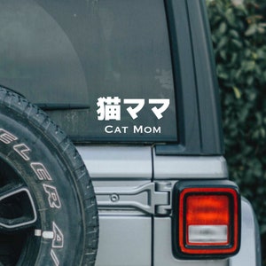 May include: A white vinyl sticker on a silver car window. The sticker says "猫ママ" in Japanese characters and "CAT MOM" in English.