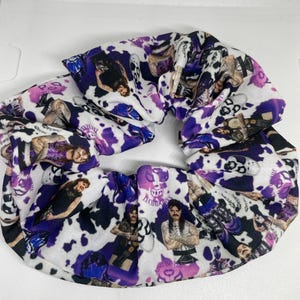 May include: A fabric hair scrunchie featuring a repeating pattern of professional wrestlers and purple cow print spots. The design includes images of wrestlers in various poses, some wearing masks, with the text Luchadores and King of the Ring.