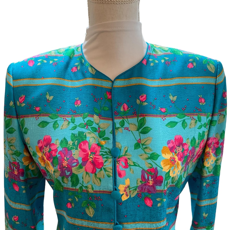 May include: A teal blazer with a floral pattern featuring pink, yellow, and purple flowers. The blazer has a mandarin collar and teal buttons. Horizontal gold stripes accent the teal fabric.