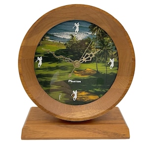 May include: A wooden desk clock with a round face featuring a golf course scene. The clock has a light brown wooden frame and base. The clock face has a green golf course with palm trees and golfers. The word "Phantom" is printed on the clock face.