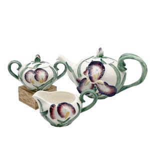 Può includere: Un servizio da tè in ceramica bianca con teiera, zuccheriera e lattiera. Ogni pezzo è decorato con un grande motivo floreale di iris nei toni del viola, rosso e giallo, con accenti verdi. La teiera ha un manico e un beccuccio curvi.