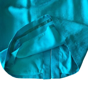 May include: A turquoise skirt with a textured fabric and a pleated design. The skirt has a matching turquoise lining and a dark green trim along the waistband. The skirt is a solid color and appears to be made of a woven material.