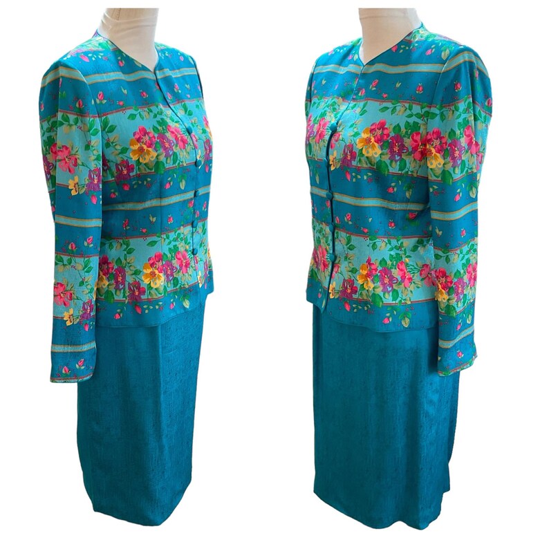 May include: A two-piece teal suit featuring a floral print jacket and a matching skirt. The jacket has a V-neck, long sleeves, and a button-down front. The floral pattern includes pink, yellow, and purple flowers. The skirt is a solid teal color.