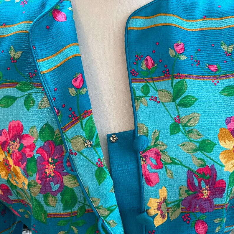 May include: A close-up of a teal floral print jacket. The jacket features a vibrant pattern of pink, purple, and yellow flowers with green leaves. The jacket has a button closure and a contrasting teal trim.