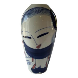 May include: A white ceramic vase with a painted face design. The vase features a woman's face with closed eyes, red lips, and a dark blue scarf. The design includes blue and red accents, creating a unique decorative piece.