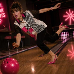 May include: A woman bowling, wearing a black and white checkered shirt with pink accents, black trousers, and pink heels. The bowling ball is red. The neon sign reads "Bowl Break Hearts" with a heart symbol.