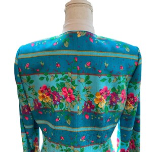 May include: A teal blazer with a floral pattern. The blazer features a vibrant design of pink, yellow, and purple flowers with green leaves. Horizontal stripes in gold and teal accent the floral print. The blazer is displayed on a mannequin.