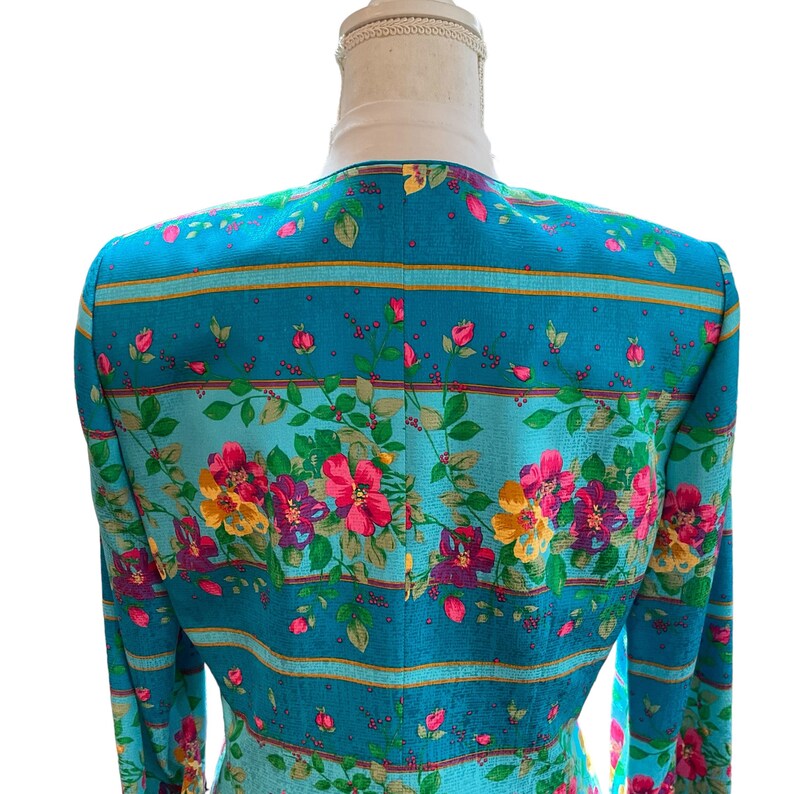 May include: A teal blazer with a floral pattern. The blazer features a vibrant design of pink, yellow, and purple flowers with green leaves. Horizontal stripes in gold and teal accent the floral print. The blazer is displayed on a mannequin.
