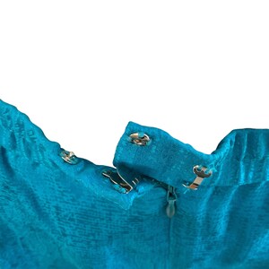 May include: Close-up of a turquoise fabric with a textured pattern. The image shows the top of a garment with metal hook-and-eye closures and a zipper. The fabric has a subtle sheen.