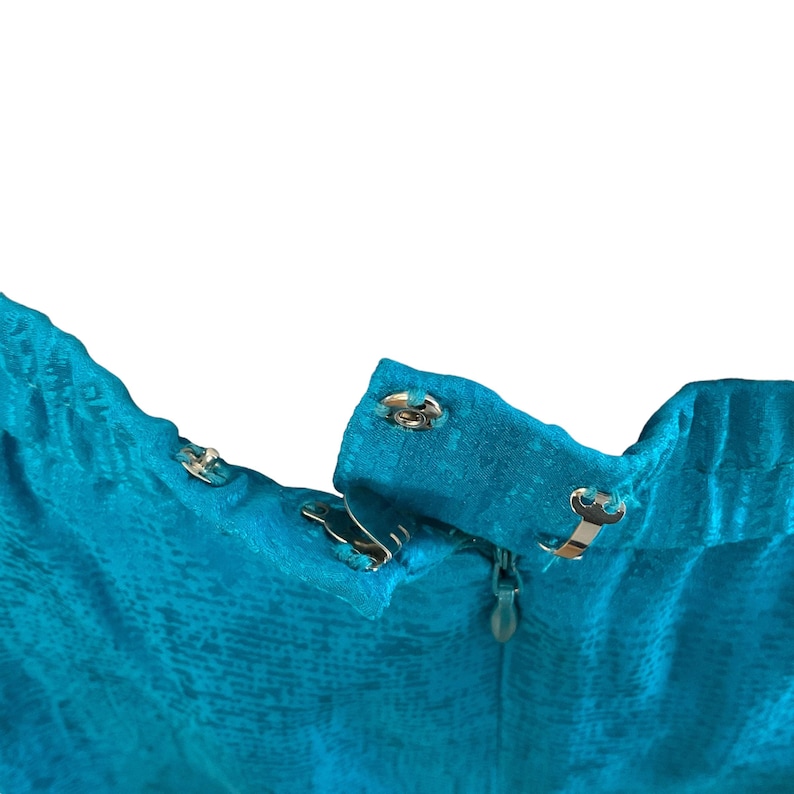 May include: Close-up of a turquoise fabric with a textured pattern. The image shows the top of a garment with metal hook-and-eye closures and a zipper. The fabric has a subtle sheen.