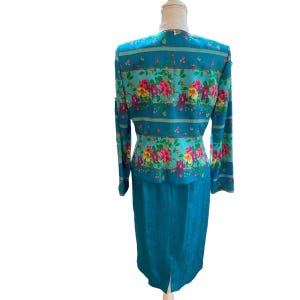 May include: A teal two-piece suit featuring a floral print jacket and a matching knee-length skirt. The jacket has long sleeves and a button-up front. The floral pattern includes pink, yellow, and red flowers.