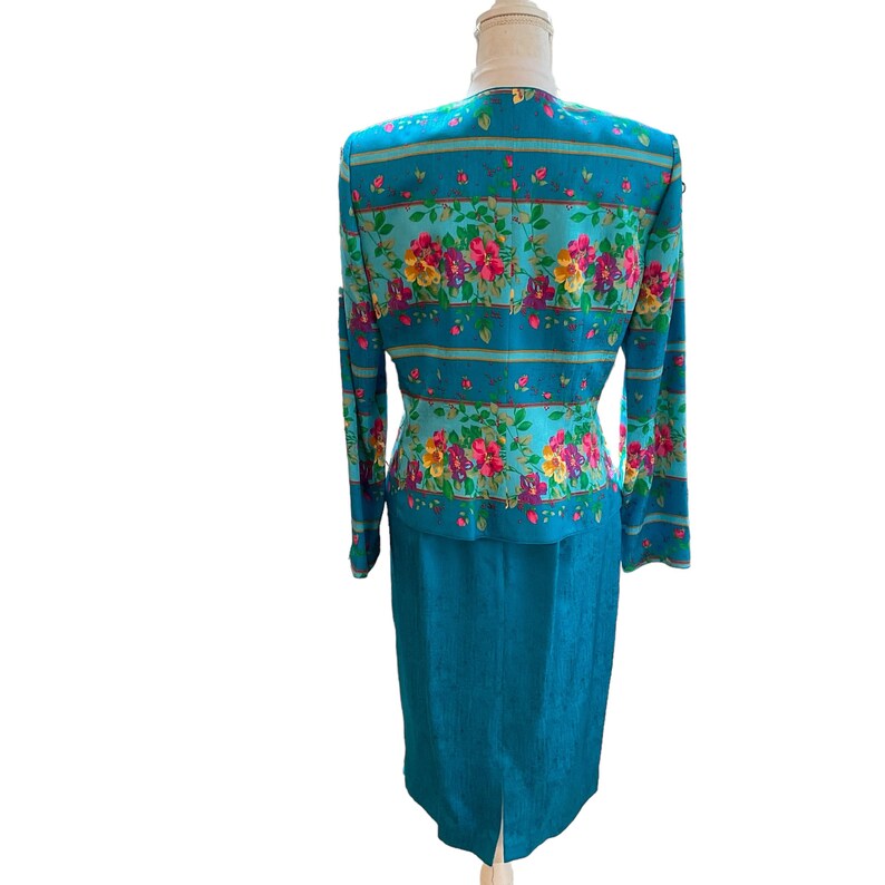 May include: A teal two-piece suit featuring a floral print jacket and a matching knee-length skirt. The jacket has long sleeves and a button-up front. The floral pattern includes pink, yellow, and red flowers.