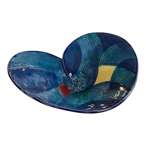 Puede incluir: Un plato de cerámica en forma de corazón hecho a mano con un acabado brillante. El plato presenta un mosaico de patrones geométricos azules, turquesas y amarillos. Un pequeño acento rojo es visible cerca del centro. Ideal para servir aperitivos o como pieza decorativa.