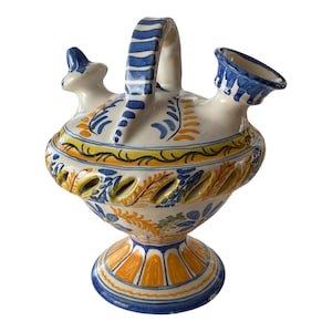 May include: A ceramic pitcher with a handle and two spouts. The pitcher is white with blue, yellow, and orange painted floral designs. The handle and spout rims are blue. The base has an orange and blue pattern.