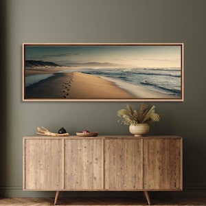 May include: A framed panoramic canvas print of a beach scene. The artwork depicts a sandy beach with footprints leading towards the ocean. The sky transitions from a light blue to a warm, golden hue, with mountains in the distance.