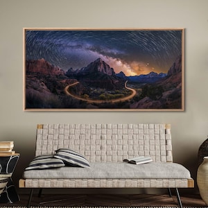 May include: A framed landscape photograph depicts a winding road illuminated by lights, snaking through a canyon under a starry night sky. The artwork features a wooden frame and is displayed above a woven sofa with striped pillows.