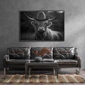 May include: Black and white photograph of a Highland cow wearing a cowboy hat. The cow is looking directly at the camera. The photo is framed and hanging on a wall above a black leather sofa.