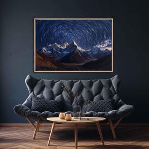 May include: A framed landscape art print featuring a mountain range under a swirling star-filled night sky. The artwork is displayed above a gray tufted sofa with decorative pillows and a wooden coffee table.