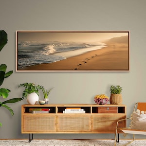 May include: A framed panoramic photograph of a beach scene. The image captures the ocean waves crashing on the shore, with footprints in the sand leading towards the horizon. The sky is a warm, golden color.