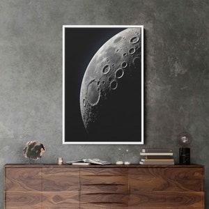 May include: A black and white photograph of the moon in a white frame. The moon is a crescent shape and shows the craters on its surface.