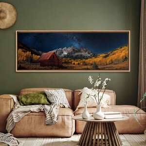 May include: A panoramic photograph of a red barn in a valley with snow-capped mountains in the background. The sky is dark blue with a bright milky way galaxy visible. The trees in the valley are a mix of green and yellow.