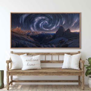 May include: A framed landscape print featuring a swirling star trail over a mountain range. The artwork has a wooden frame and is displayed above a wooden bench with pillows. One pillow reads "welcome home".