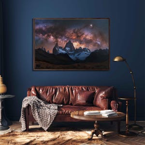 May include: A framed photograph of a mountain range with a starry sky above. The mountains are snow-capped and the sky is a deep blue with white clouds. The photo is hanging on a blue wall above a brown leather couch.