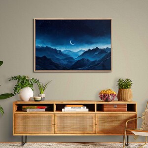May include: A framed landscape painting depicts a crescent moon and stars in a dark blue night sky above blue mountain ranges. The artwork is displayed above a wooden cabinet with woven doors, holding plants and decorative items.