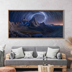 May include: A panoramic photograph of a mountain range under a starry night sky. The stars are captured as streaks of light, creating a swirling pattern in the sky. The mountains are silhouetted against the dark sky, with a few peaks illuminated by the moonlight.