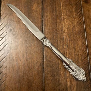 May include: A silver butter knife with an ornate handle. The blade is long and thin, and the handle is decorated with intricate details.