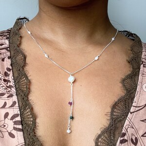 Op de afbeelding: Een zilveren ketting met een grote witte parel hanger en een kleinere parel hanger met groene en roze edelstenen.