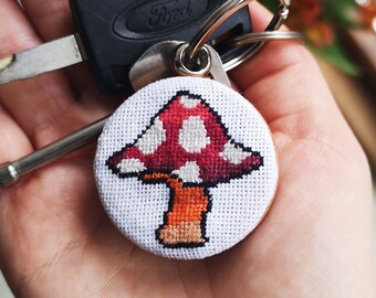 Puede incluir: Un llavero redondo de punto de cruz blanco con un hongo rojo con puntos blancos y un tallo naranja. El llavero está unido a un juego de llaves, incluida una llave de coche Ford. El fondo incluye una ventana y flores naranjas.