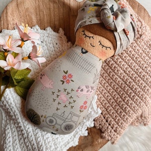 以下が含まれることがあります： 茶色の顔、閉じられた目、そしてピンクの頬をした手作り布人形。人形は、花柄のグレーと白の布のドレスと、花柄のグレーと白の布のターバンを着ています。人形は、白い編み物の毛布とピンクの編み物の毛布の上に横たわっています。