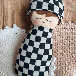 以下が含まれることがあります： 黒と白のチェック柄の布製のドールで、茶色の髪と目を閉じている。ドールは黒と白のチェック柄の帽子をかぶっており、白い編み物の毛布の上に横たわっている。
