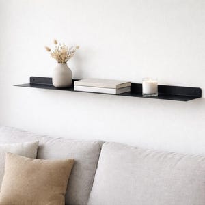 Könnte beinhalten: Ein schwarzes schwebendes Metallregal an einer weißen Wand, das eine beige Vase mit Trockenblumen, zwei Bücher und eine brennende Kerze in einem Glas trägt. Darunter befindet sich ein helles Sofa mit Kissen.