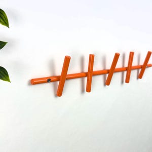 Könnte beinhalten: Wandgarderobe aus orangefarbenem Holz mit sechs Haken. Die Haken sind abgewinkelt und gleichmäßig auf einer horizontalen Stange angeordnet. Die Garderobe ist an einer weißen Wand befestigt, mit grünen Blättern in der Ecke.