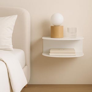 Könnte beinhalten: Minimalistische Schlafzene mit einem weißen, schwebenden Nachttischregal mit einer kleinen Lampe, einem Glas und Büchern. Links ein Bett mit weißem Kissen und Bettwäsche. Die Farbpalette ist neutral mit Beige- und Weißtönen.