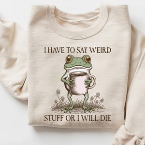 May include: Cream-colored sweatshirt with the text "I HAVE TO SAY WEIRD STUFF OR I WILL DIE." A cartoon frog holding a coffee mug is printed on the front. The frog is green with brown eyes and the mug is pink.