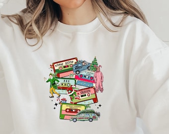 May include: White sweatshirt with a colorful graphic of cassette tapes featuring characters from popular Christmas movies, including "Home Alone", "Elf", "The Grinch", and "A Christmas Story".