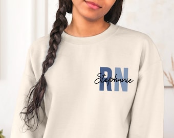 May include: A white sweatshirt with a blue and white gradient "RN" design and the name "Stephanie" written in cursive below.