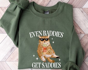 Peut inclure: Sweat-shirt vert avec un chat de dessin animé portant des lunettes de soleil et tenant un seau de pop-corn. Le texte "Even Baddies Get Saddies" est imprimé sur le sweat-shirt.