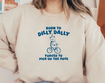 May include: A cream-colored sweatshirt with a blue graphic of a bear riding a bicycle. The graphic says "Born to Dilly Dally, Forced to Pick Up the Pace."