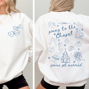 May include: White sweatshirt with blue graphic text and illustrations. The front features two heart-shaped pendants with the text "Mrs. Wilson". The back displays the phrase "going to the Chapel gonna get married" with wedding-themed drawings.