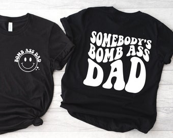 May include: Two black t-shirts with white text. One shirt says "Somebody's Bomb Ass Dad" in a retro font. The other shirt has a smiley face and the text "Bomb Ass Dad".