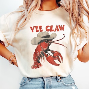 May include: Cream-colored t-shirt featuring a red lobster wearing a cowboy hat. The text "Yee Claw" is printed above the lobster. The design has a vintage, slightly distressed look.