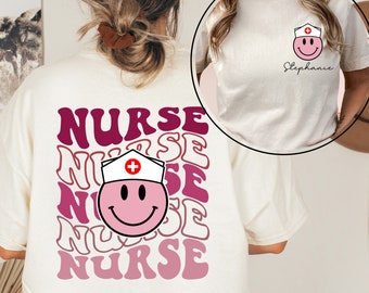 May include: Cream-colored t-shirt featuring a retro design with the word "NURSE" stacked multiple times in shades of pink and burgundy. A smiling face wearing a nurse's cap is incorporated into the design. A smaller version of the design is on the front.