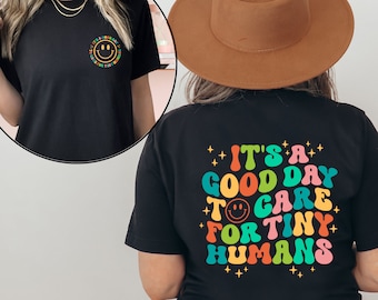 May include: Black t-shirt with the text "It's a good day to care for tiny humans" in colorful, retro-style lettering. The shirt also features a smiley face graphic and stars. A smaller version of the design is on the front.